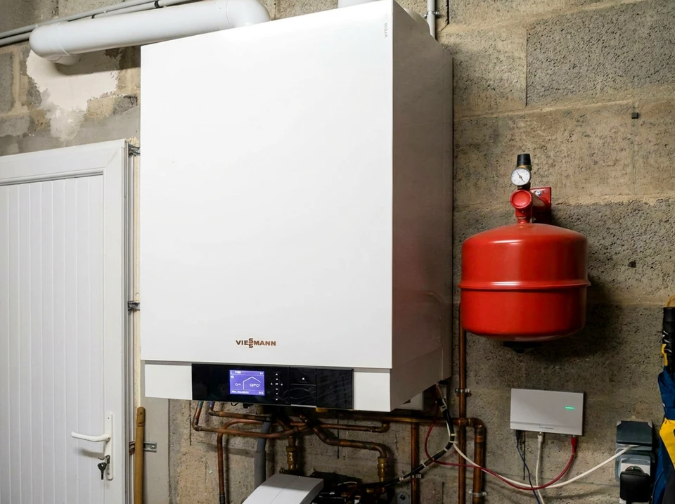Pompe à chaleur air/eau installée en extérieur, fixée sur un châssis métallique réglable, positionnée contre un mur en pierre apparente entre deux volets en bois peints en rouge